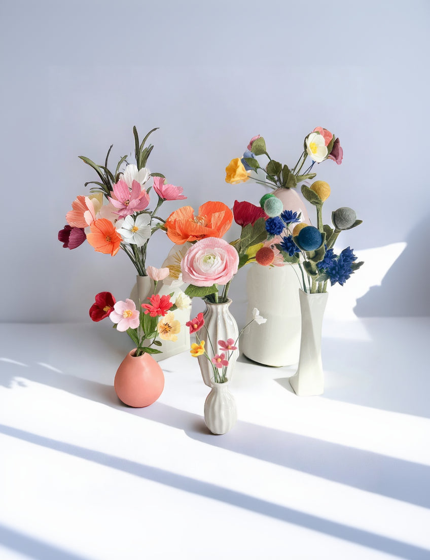 Three ceramic vases in white, peach, and ribbed white styles, each holding a bouquet of artificial multicolored flowers, arranged on a white surface against a pale blue-gray background.