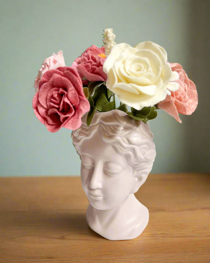 Alt text: A classical bust-shaped vase featuring a serene face, holding a felt flower bouquet with pink, cream, and red roses. The soft pastel colors of the flowers add a gentle contrast to the minimalist white surface and background, creating an elegant display.