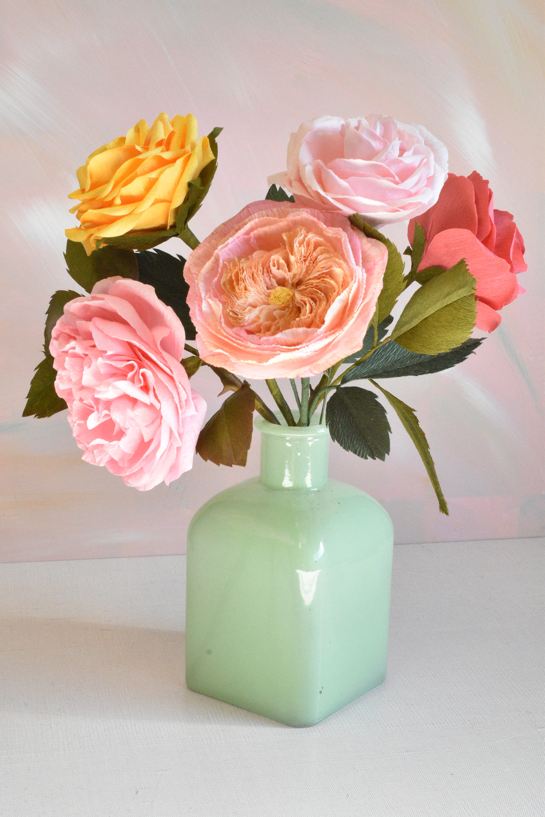 A bouquet of artificial roses in pink, peach, yellow, and coral, arranged in a glossy pale green glass vase against a soft pink and white background.