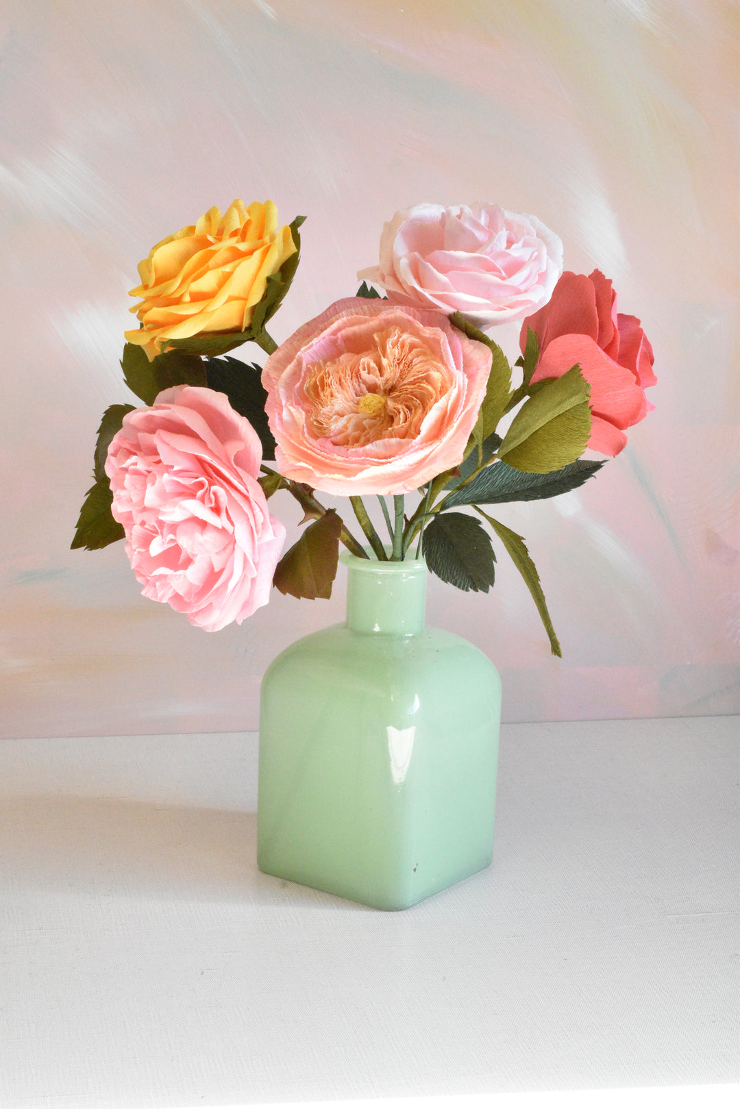 A bouquet of artificial roses in pink, peach, coral, and yellow, arranged in a small mint green glass vase on a white surface against a soft pastel background.