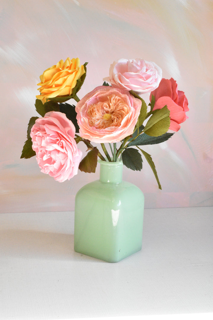 A bouquet of artificial roses in pink, peach, coral, and yellow, arranged in a small mint green glass vase on a white surface against a soft pastel background.
