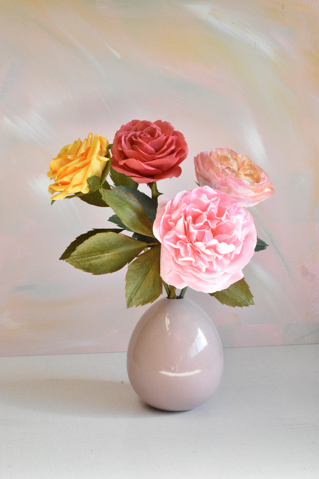 A ceramic egg-shaped vase in a pale pinkish-gray color, holding a bouquet of artificial flowers including a large pink rose, a red rose, a yellow rose, and a peach-colored rose with green leaves, set against a soft pastel painted background.