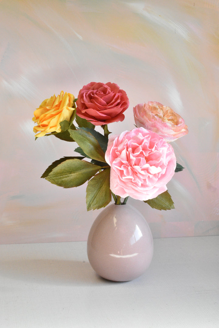 A ceramic egg-shaped vase in a pale pinkish-gray color, holding a bouquet of artificial flowers including a large pink rose, a red rose, a yellow rose, and a peach-colored rose with green leaves, set against a soft pastel painted background.