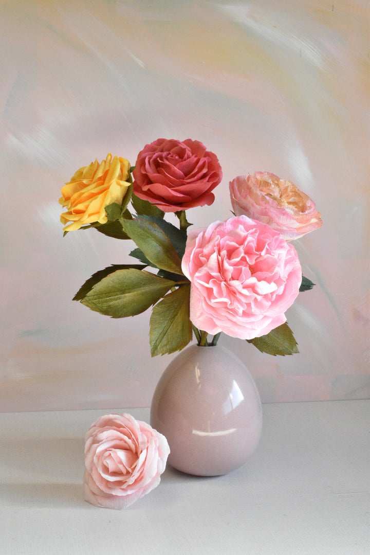 A bouquet of artificial roses in pink, red, yellow, and peach tones arranged in a small pale pink vase, with a matching pink rose placed in front on a light surface against a soft painted pastel background.