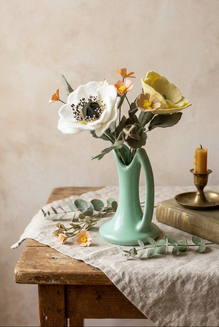 A small mint green ceramic vase holding a bouquet of artificial flowers, including a large white anemone with a black center, a pale yellow poppy, and smaller orange and cream blossoms, set against a soft light pink background.