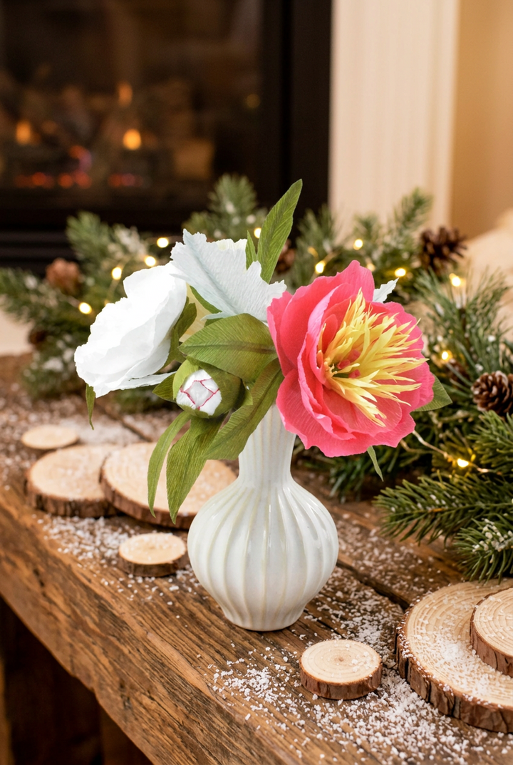 A white ribbed ceramic vase holding a faux floral arrangement with a large pink flower, a white flower, and green leaves, styled on a rustic wooden mantel with holiday greenery and warm lights in the background.