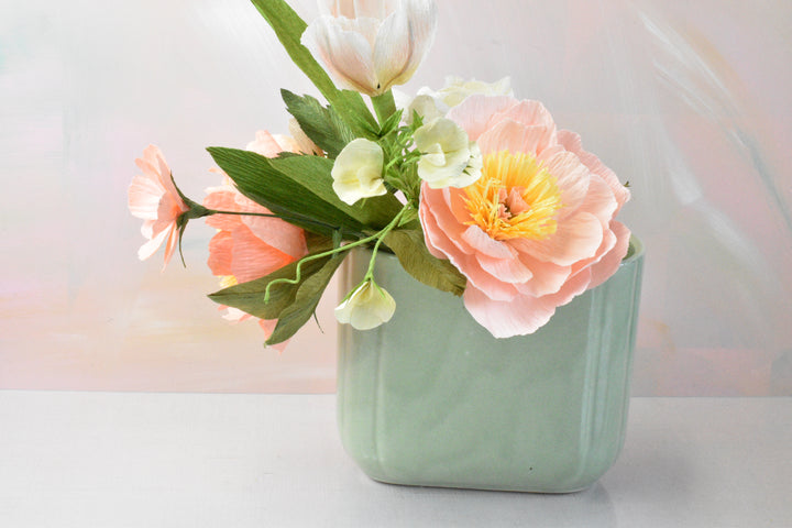 A pastel floral arrangement in a square mint-green ceramic vase, featuring a large peach-pink peony with a yellow center, white tulips, and green leaves, set against a soft, painterly pastel background.