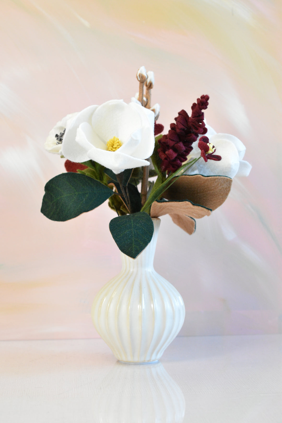 A faux floral arrangement featuring white calla lilies, dark burgundy berries, and green leaves in a ribbed white ceramic vase, set against a soft pastel pink and yellow background.