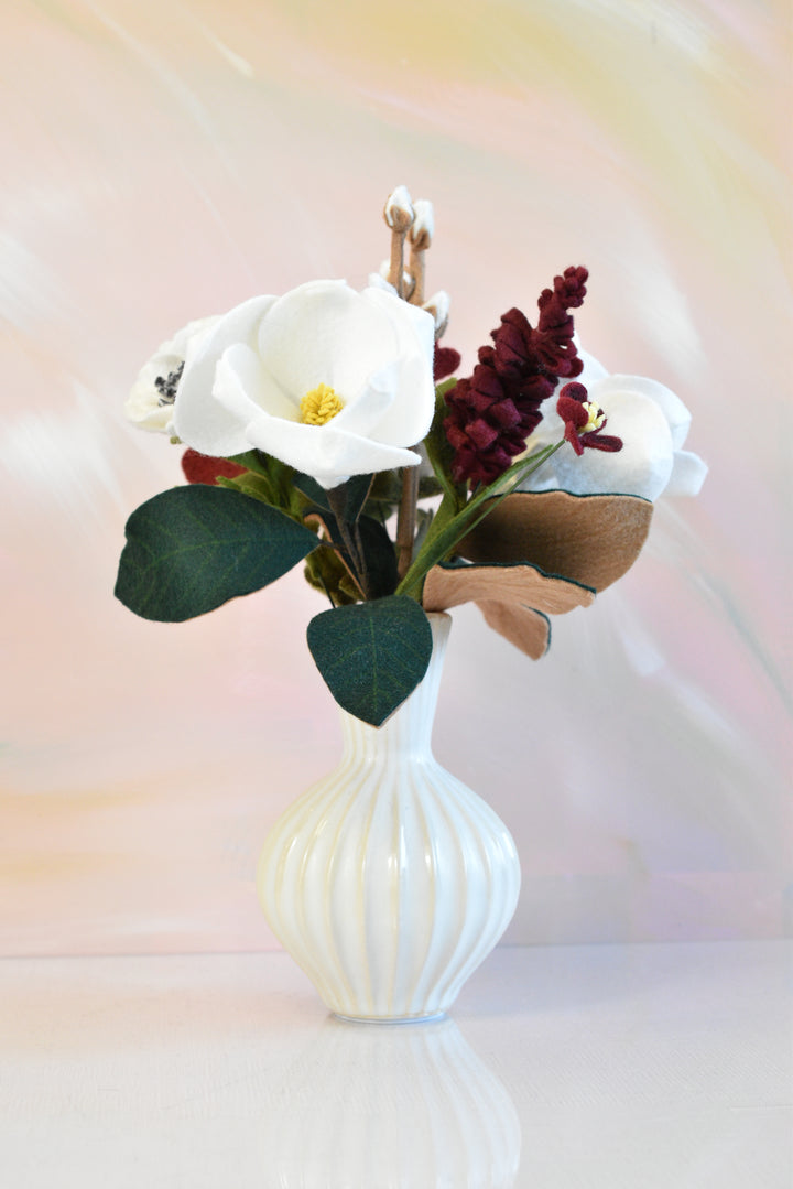 A faux floral arrangement featuring white calla lilies, dark burgundy berries, and green leaves in a ribbed white ceramic vase, set against a soft pastel pink and yellow background.