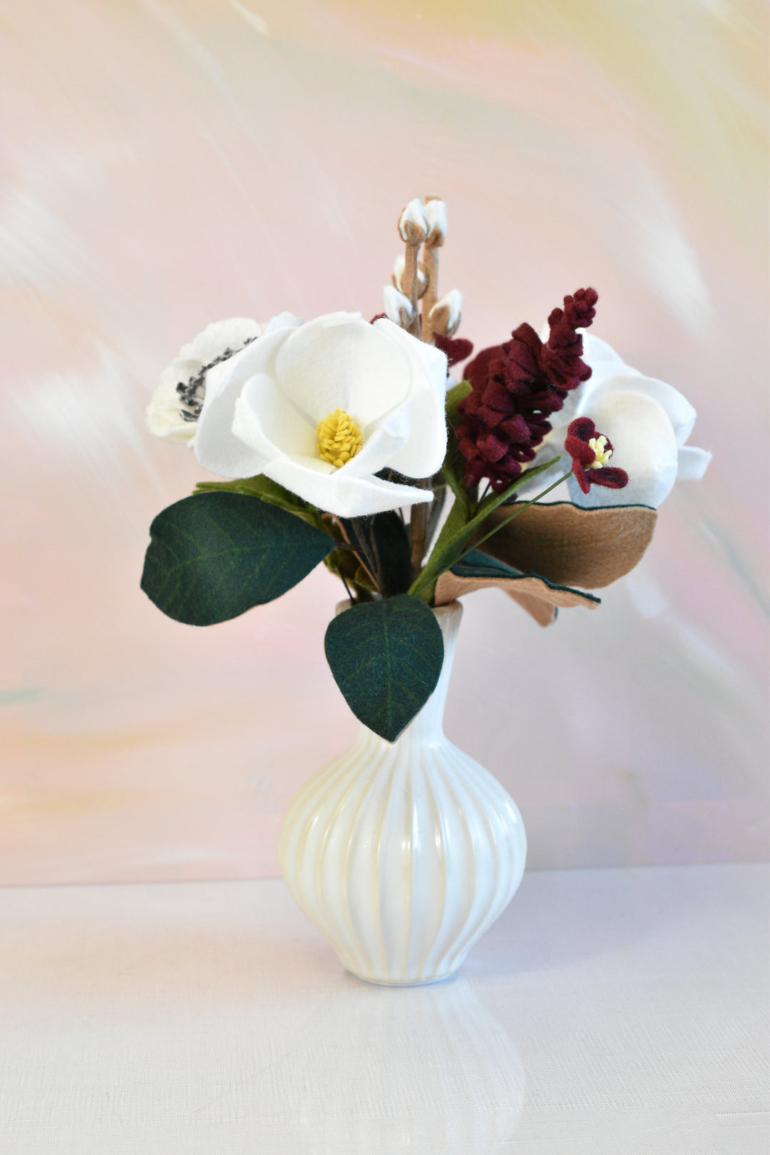 A faux floral arrangement featuring white magnolia blossoms, dark green leaves, burgundy accents, and cotton stems in a ribbed white ceramic vase, set against a soft pastel background.