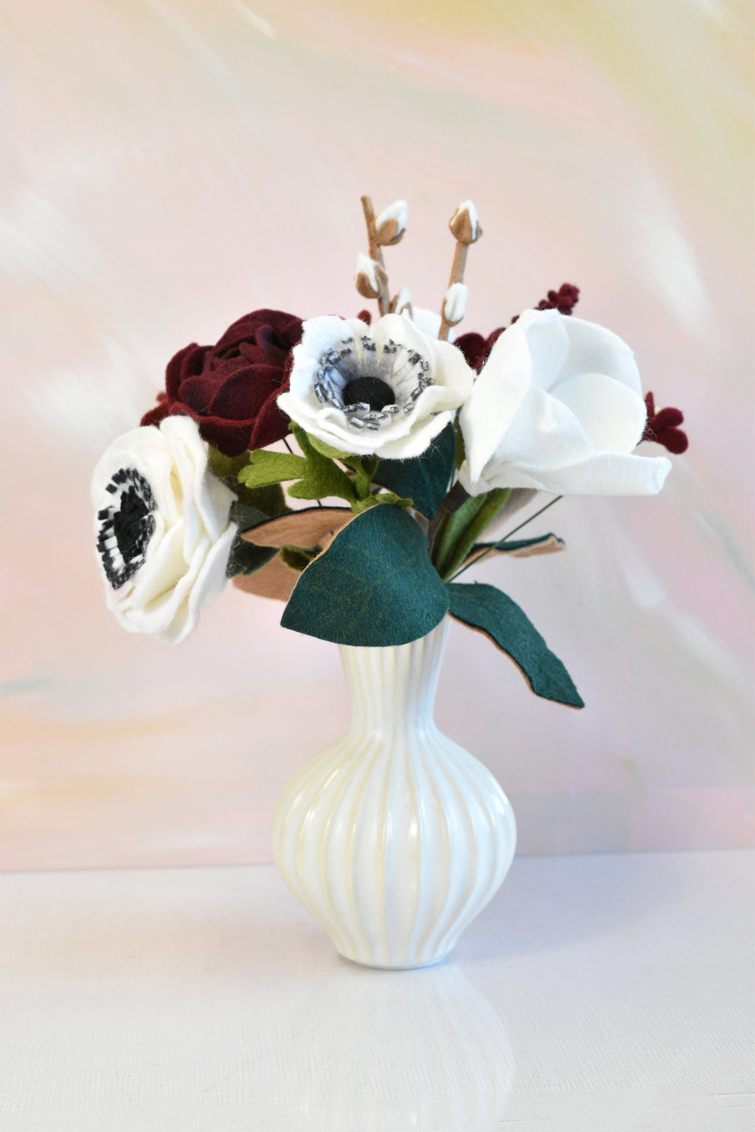 A faux flower arrangement featuring white anemone-style blooms, deep burgundy roses, and green leaves in a ribbed white ceramic vase, set against a soft pastel pink and yellow background.