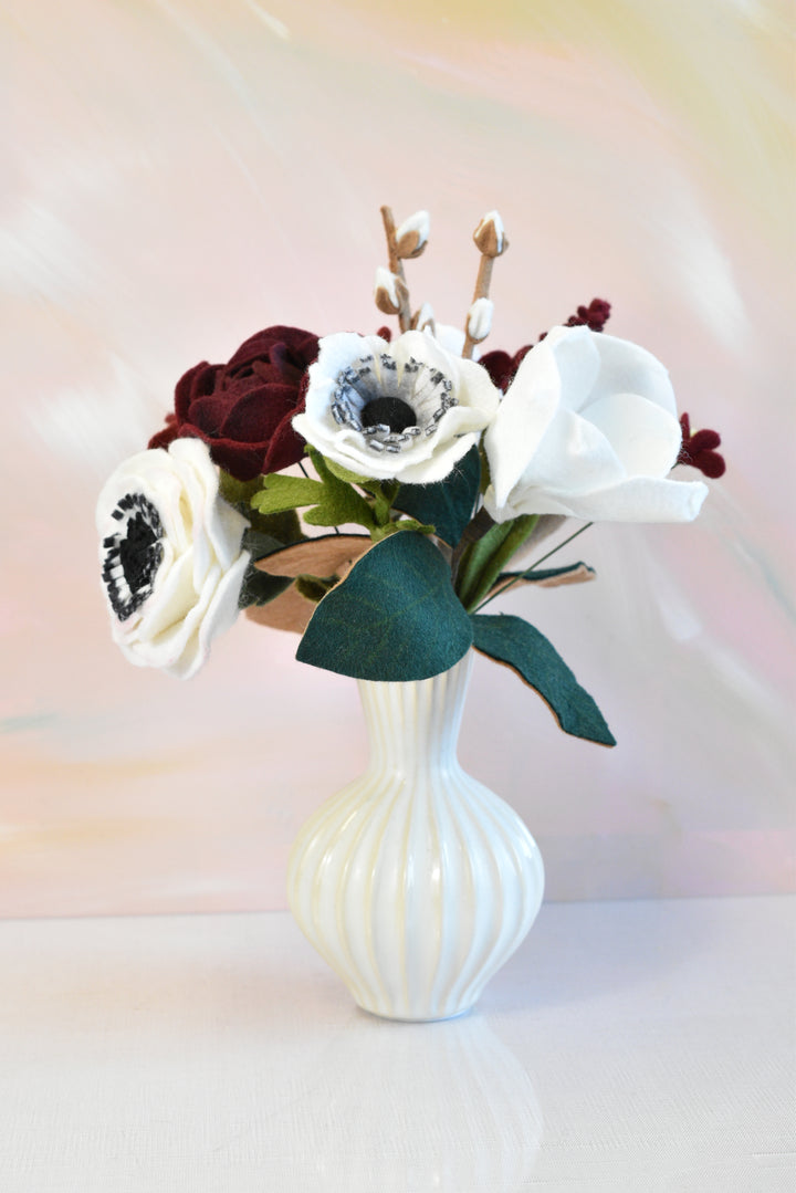 A faux flower arrangement featuring white anemone-style blooms, deep burgundy roses, and green leaves in a ribbed white ceramic vase, set against a soft pastel pink and yellow background.