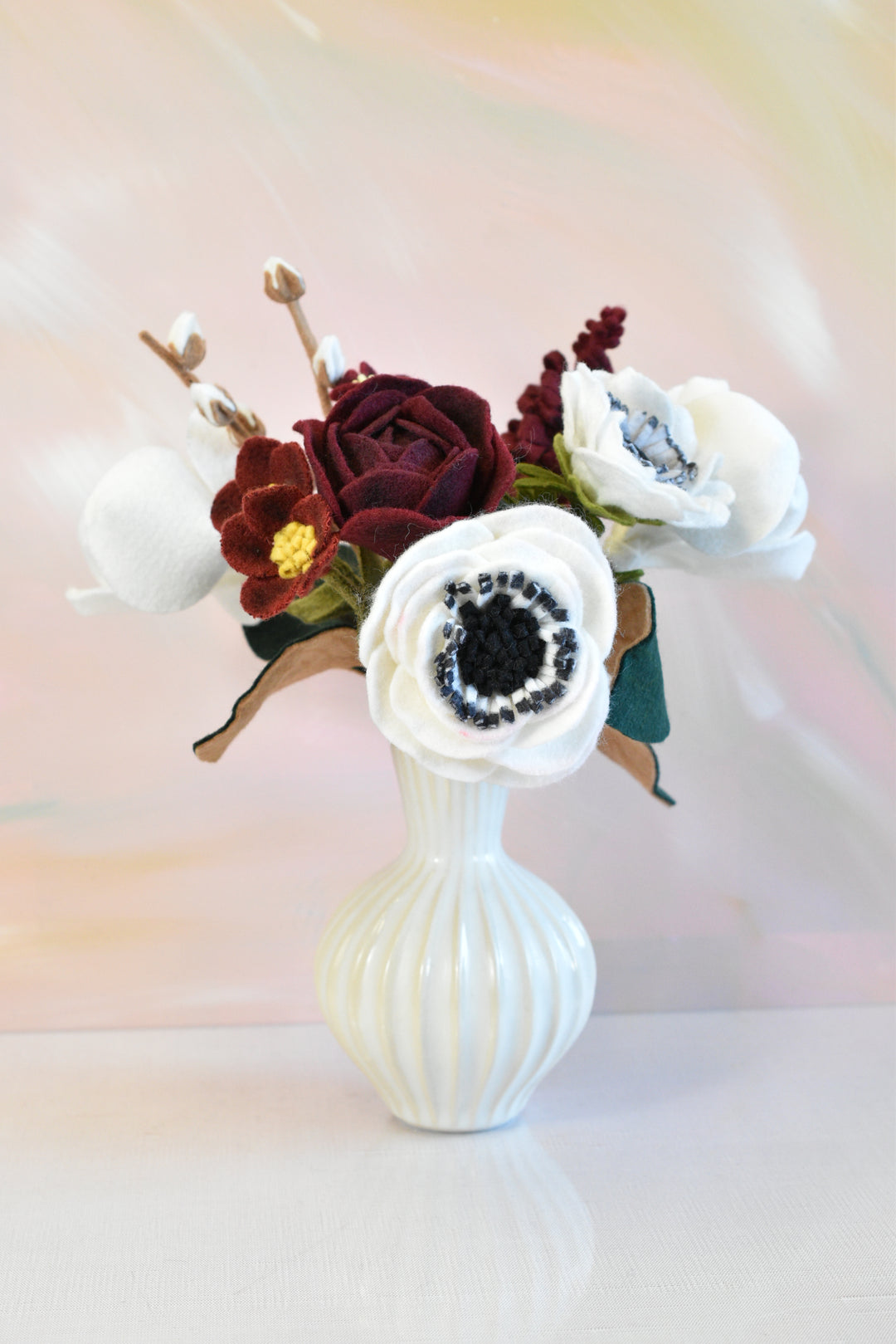 A decorative artificial flower arrangement in a small ribbed white ceramic vase, featuring white anemone-style flowers, a burgundy rose, and dark burgundy accents against a soft pastel background.