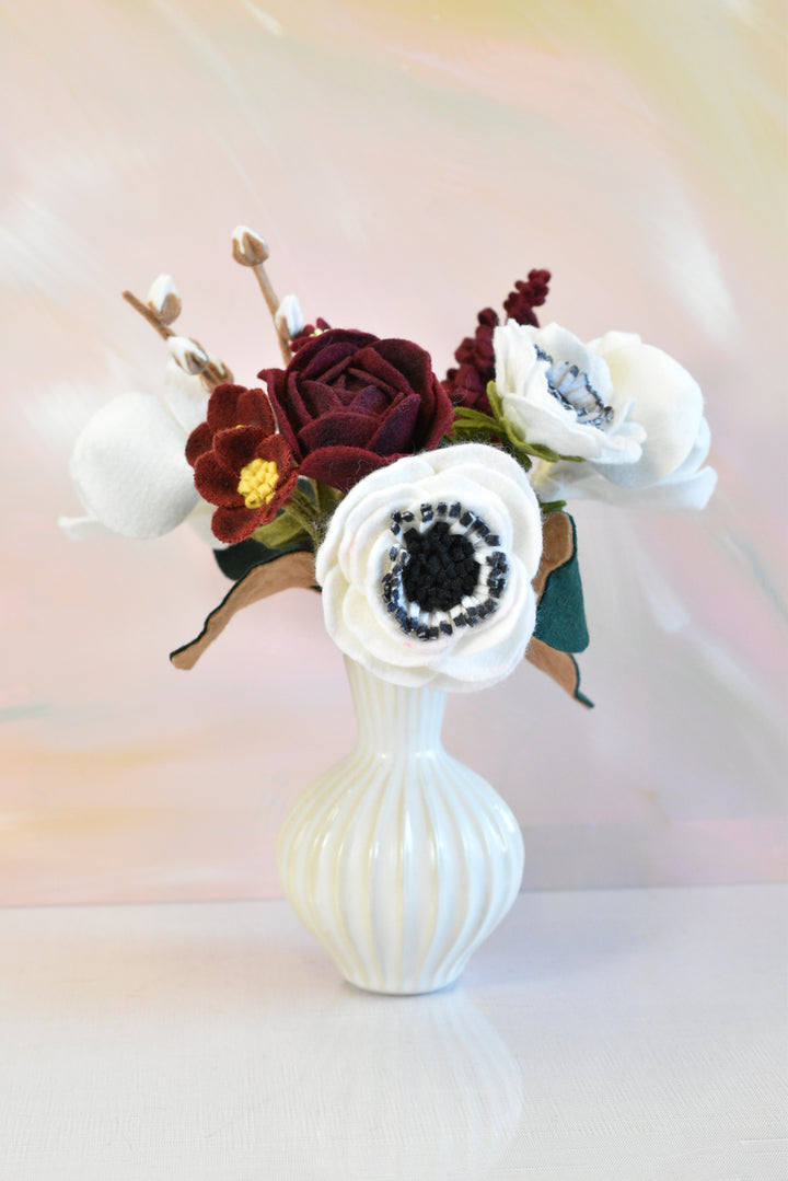 A decorative artificial flower arrangement in a small ribbed white ceramic vase, featuring white anemone-style flowers, a burgundy rose, and dark burgundy accents against a soft pastel background.