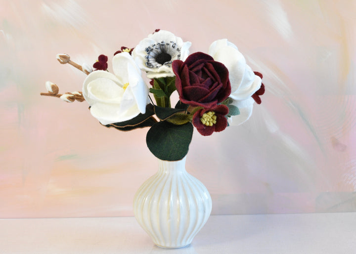 A faux floral arrangement featuring white magnolia blossoms, a deep burgundy rose, and white anemone flowers with dark green foliage, presented in a white ribbed ceramic vase on a white surface against a soft pastel background.