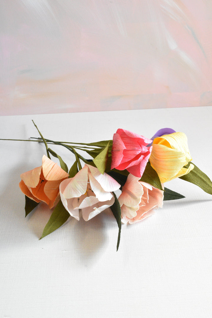 A bouquet of paper tulips with green stems and leaves, featuring pastel shades of orange, pink, peach, yellow, and purple, arranged on a white surface against a soft pink and white background.