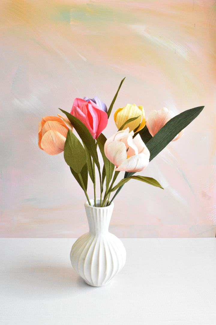 A white ribbed ceramic vase holding a bouquet of artificial tulips in pastel shades of pink, peach, yellow, and white, set against a soft pastel painted background.