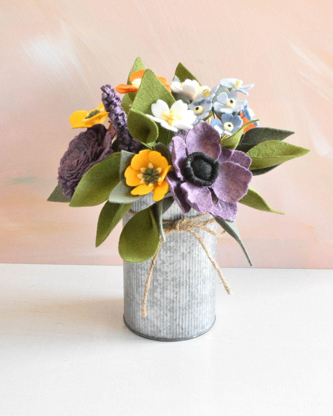 A small felt flower bouquet in a galvanized metal tin vase, featuring purple, yellow, white, and blue felt flowers with green felt leaves, set against a soft peach-pink background.