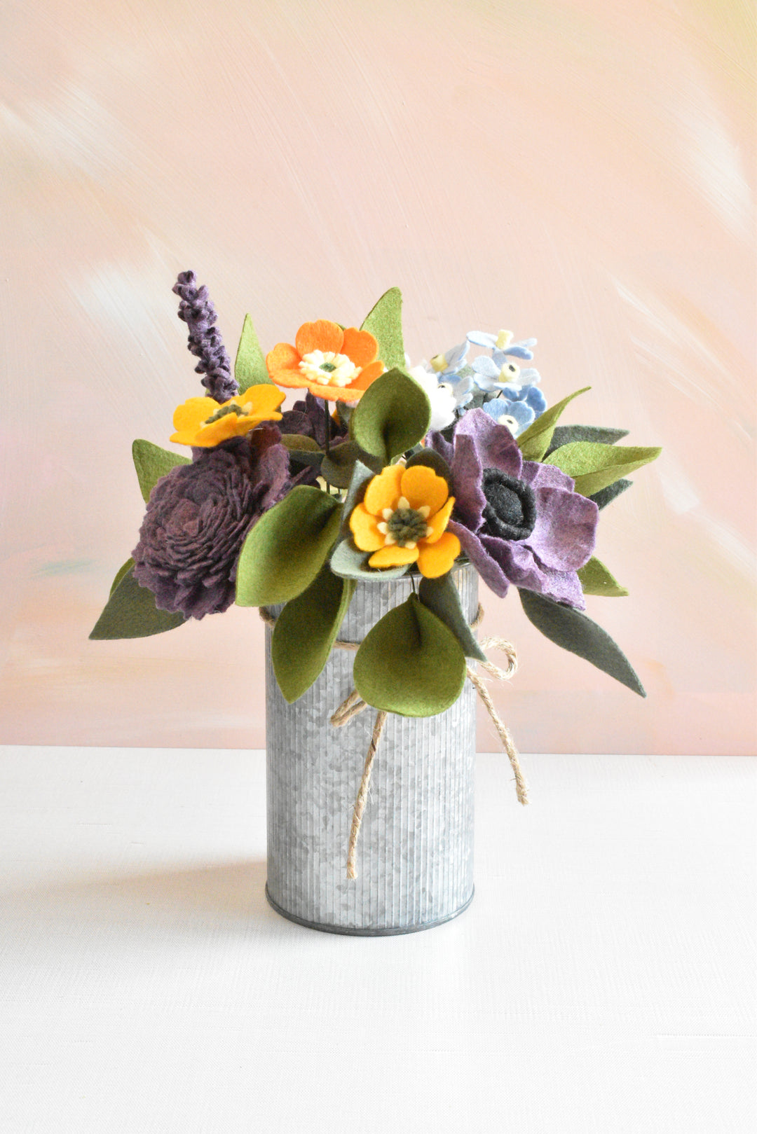 A decorative artificial flower arrangement in a galvanized metal vase, featuring orange poppy-like flowers, purple anemone-style blooms, green leaves, and a small light blue flower, set against a soft peach background.