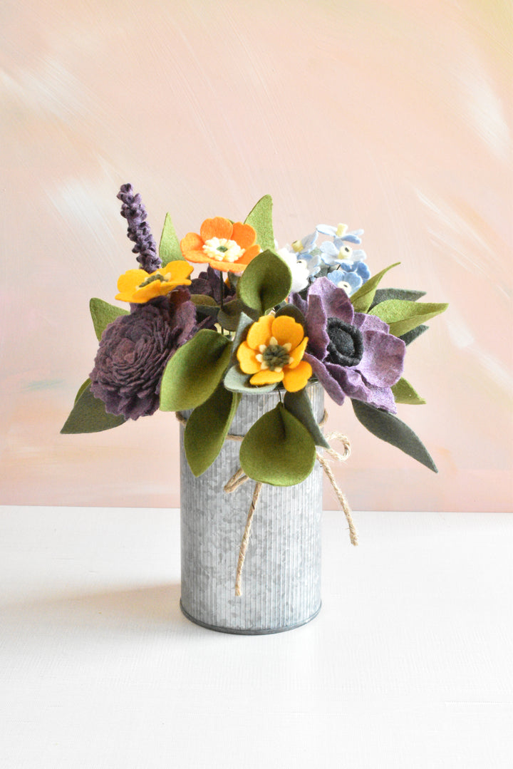 A decorative artificial flower arrangement in a galvanized metal vase, featuring orange poppy-like flowers, purple anemone-style blooms, green leaves, and a small light blue flower, set against a soft peach background.