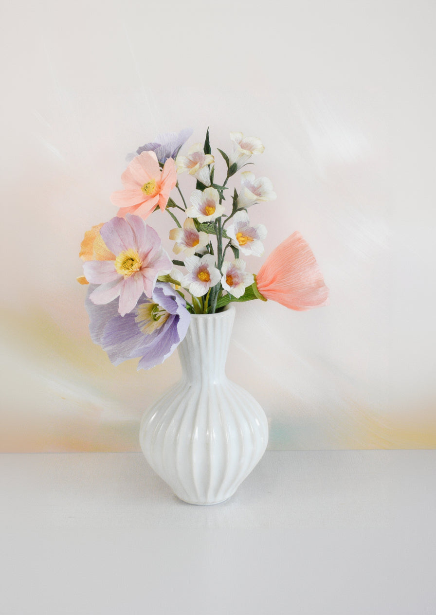 A white ribbed ceramic vase holding a bouquet of pastel artificial flowers, including lavender, peach, and white blossoms, set against a soft blurred abstract background.