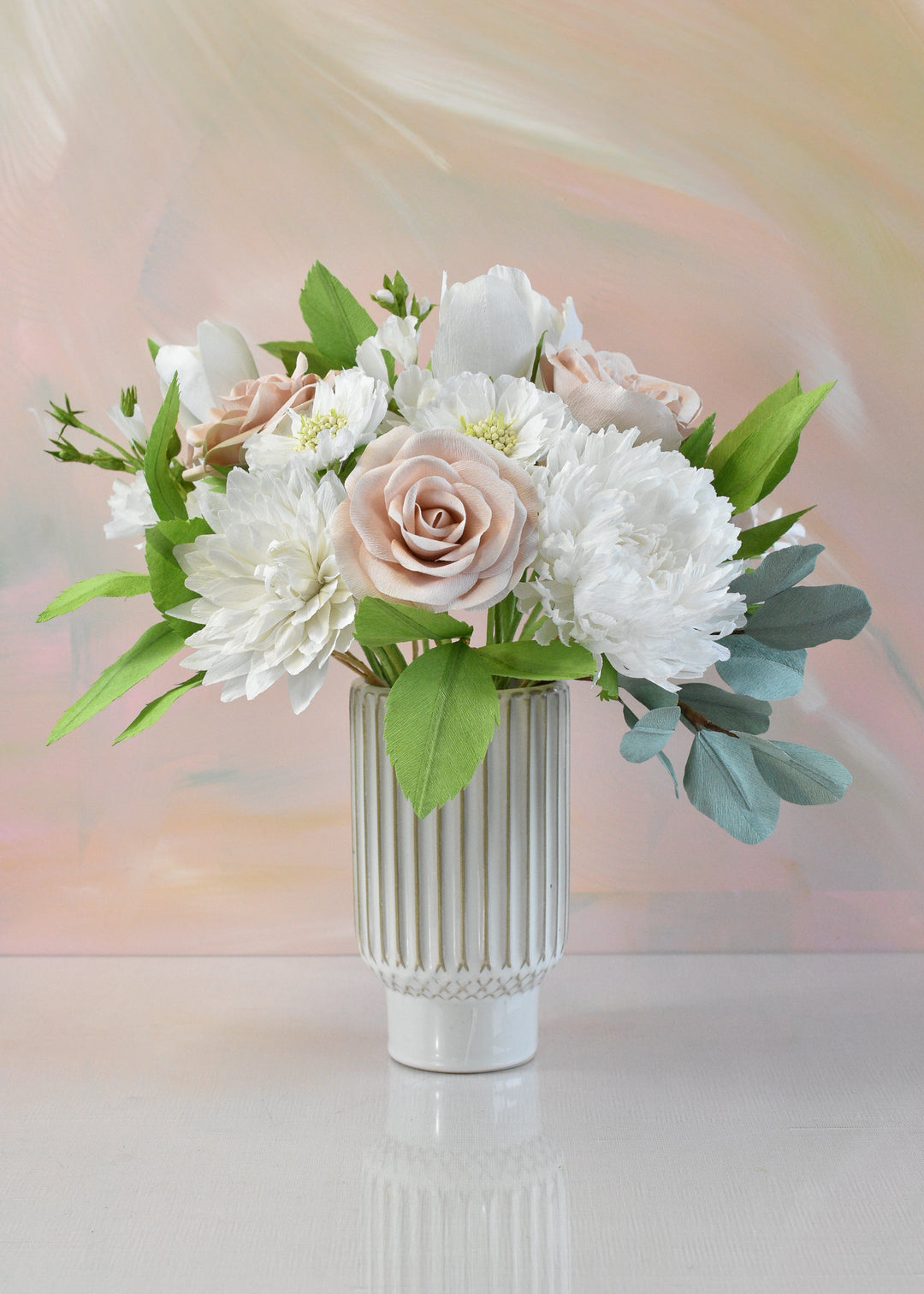 A floral arrangement featuring white and blush pink flowers with green leaves, presented in a white ribbed ceramic vase on a reflective white surface against a soft pastel background.