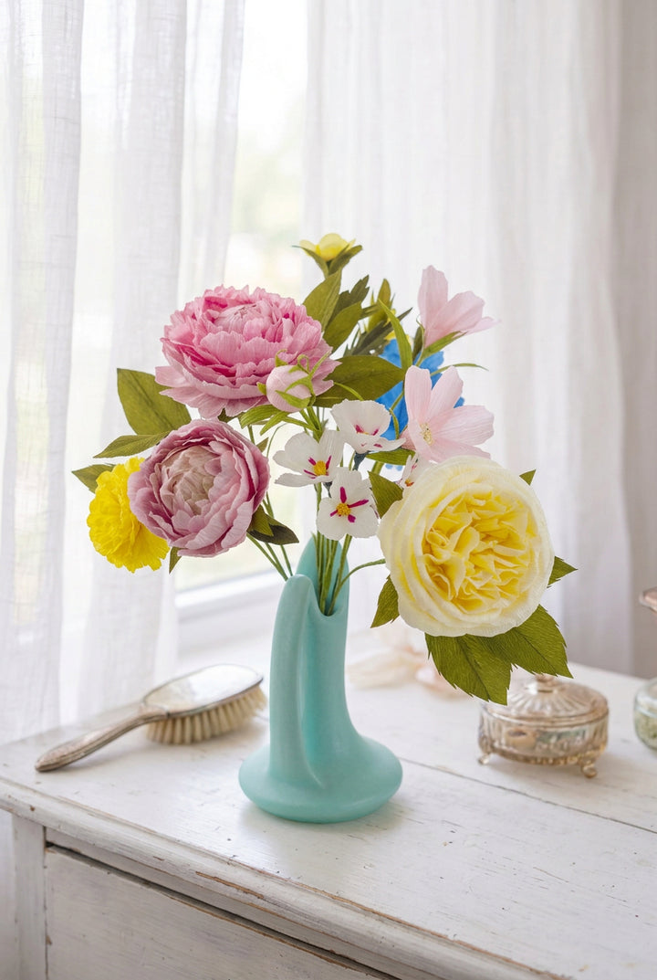 A bouquet of paper flowers in a small mint green vase, featuring pink peonies, a pale yellow rose, white blossoms with pink centers, and green leaves on a light background.