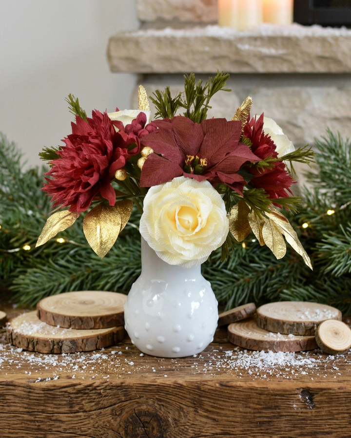 A festive holiday floral arrangement in a small white textured vase, featuring burgundy poinsettias, a cream rose, gold leaves, and green pine branches, styled on a wooden mantel with wood slices and snowflake decorations.
