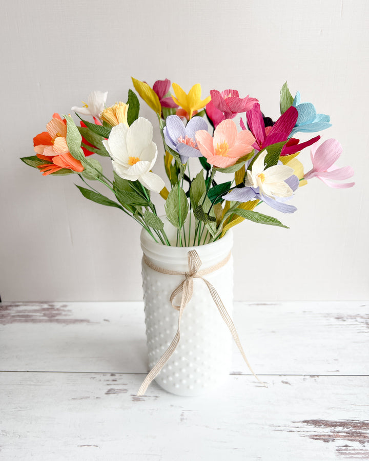 A colorful bouquet of paper wildflowers in shades of pink, yellow, orange, blue, and white, arranged in a textured white vase. The vase is decorated with a tied ribbon, adding a rustic charm. The bouquet sits on a light wooden surface against a plain background.