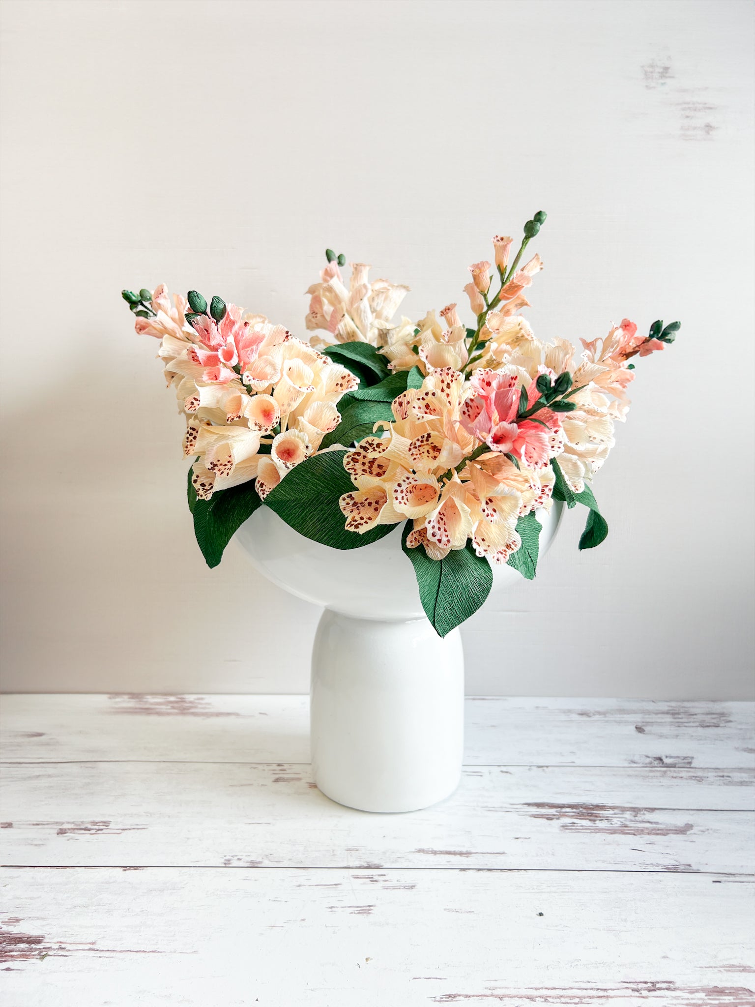A lifelike artificial foxglove stem with pink and white flowers in a white vase, placed on a wooden surface.