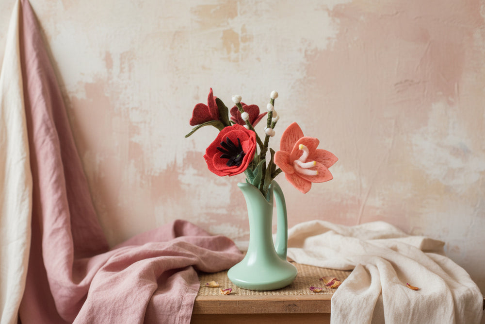 Green vase with red and pink flowers on a wooden table against a textured wall.
