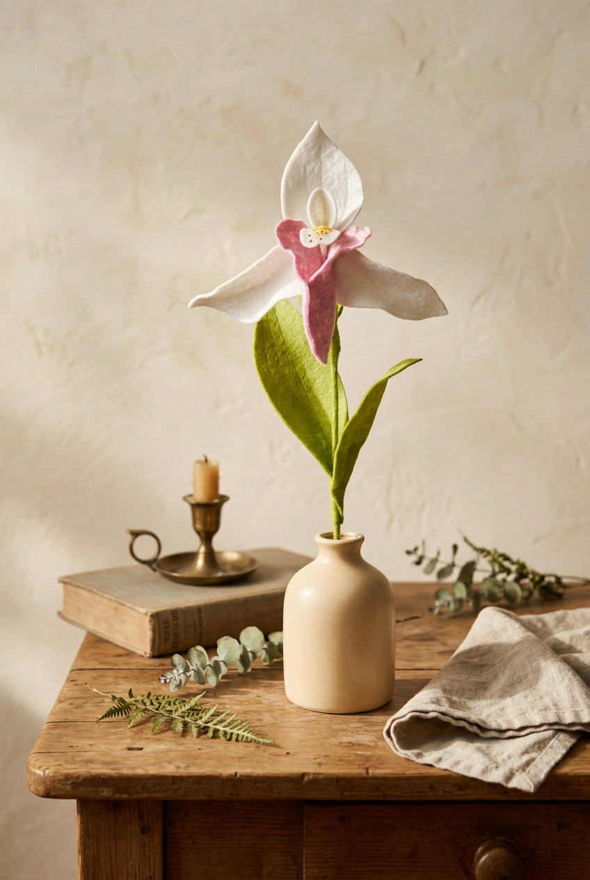 A single artificial Lady Slipper Stem with white petals, a pink center, and green leaves, standing in a small teal plastic pot on a white surface against a soft pastel painted background.