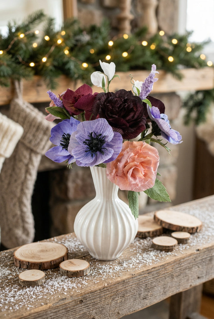 A white ribbed ceramic vase holding a mixed floral arrangement with purple anemone-like flowers, a peach rose, dark burgundy blooms, and green leaves, set against a soft pastel abstract background.