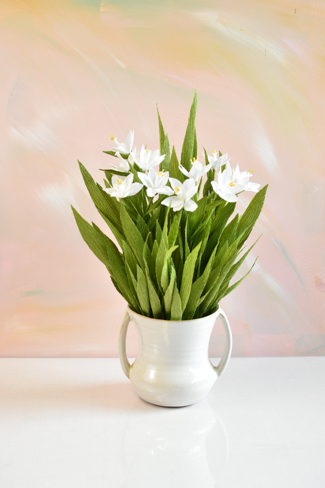 White ceramic pitcher vase filled with fresh green leaves and white flowers, photographed against a soft pastel pink and peach background.