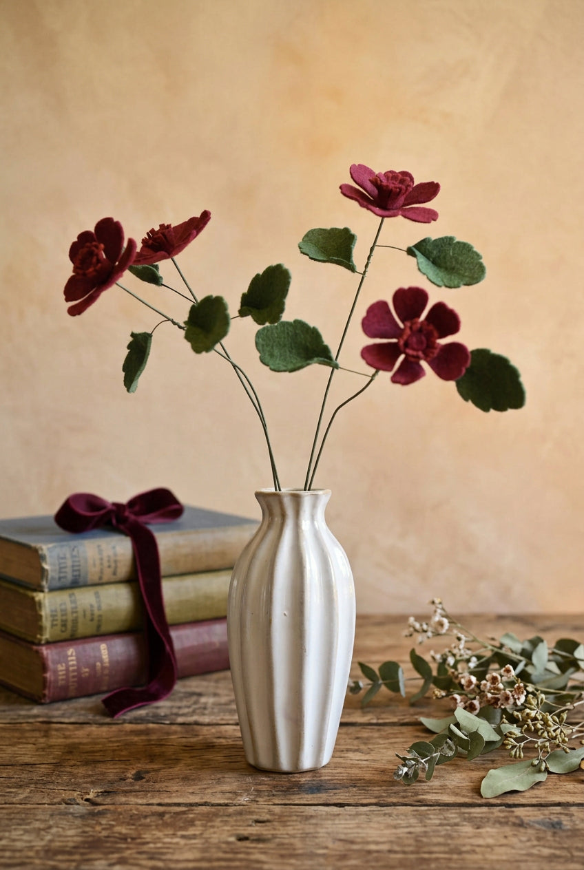 Three felt chocolate cosmos with green leaves arranged in a white ribbed ceramic vase, set against a soft pastel pink and cream painted background.