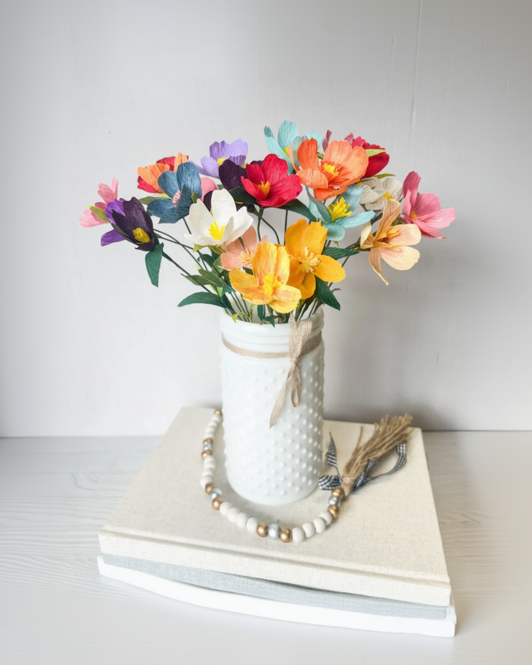A bouquet of colorful artificial flowers arranged in a white textured vase tied with a beige ribbon, placed on stacked neutral books with a beaded garland and dried grasses in front, against a soft light gray background.