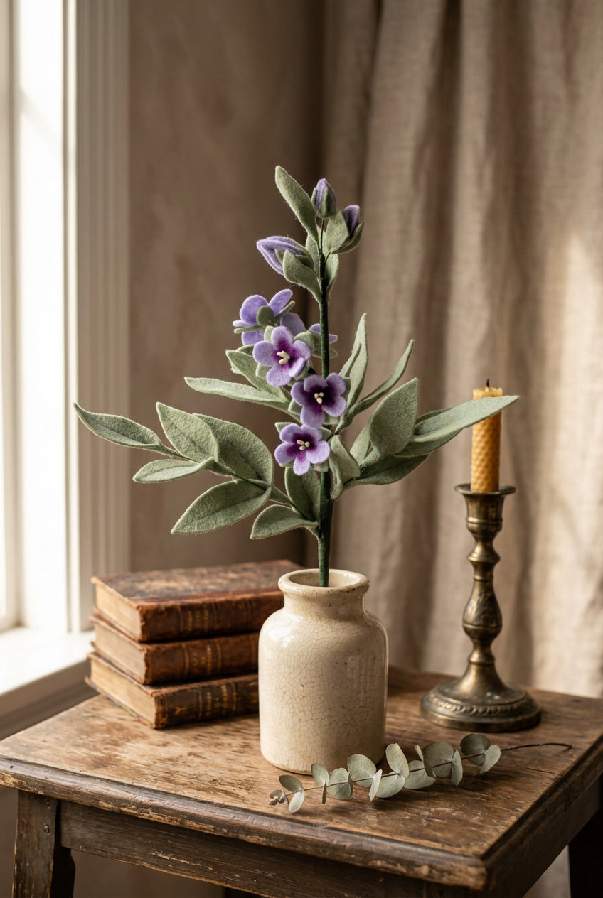 A felt Jacob's Ladder stem with purple blossoms and muted green leaves, mounted in a round green holder against a soft light beige background.