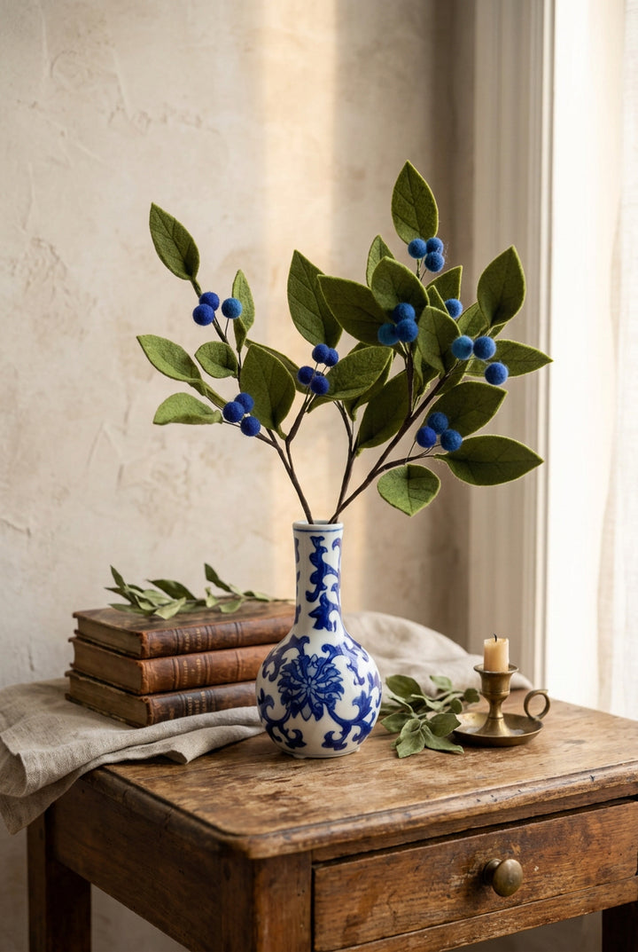 Felt berry branch with green leaves and blue berries arranged in a small blue and white patterned ceramic vase.