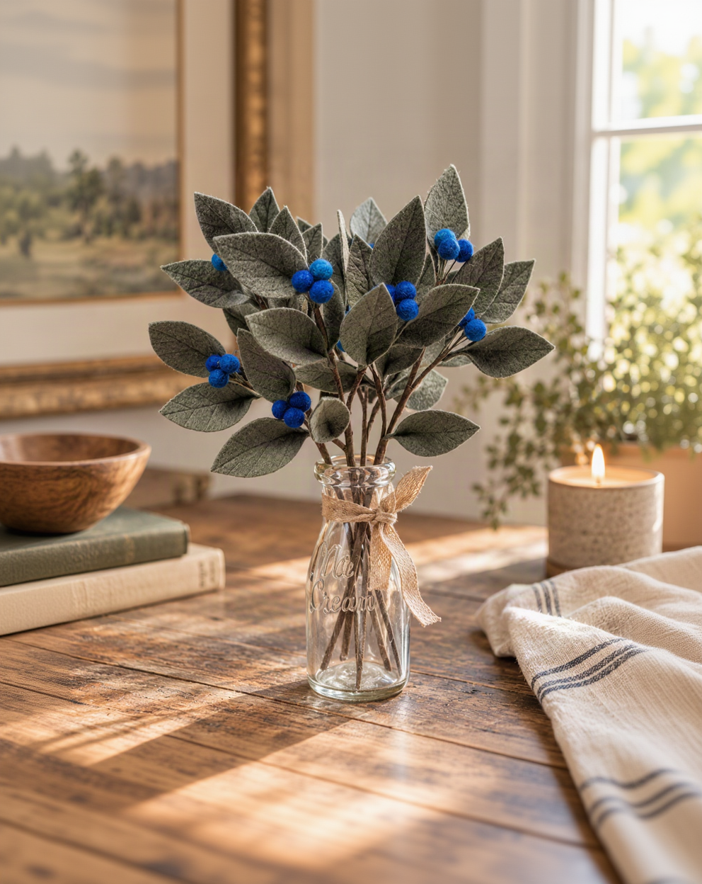 Faux blue berry branch arrangement in a clear glass jar vase with a burlap ribbon, displayed on a rustic wooden table in a warm, sunlit room.