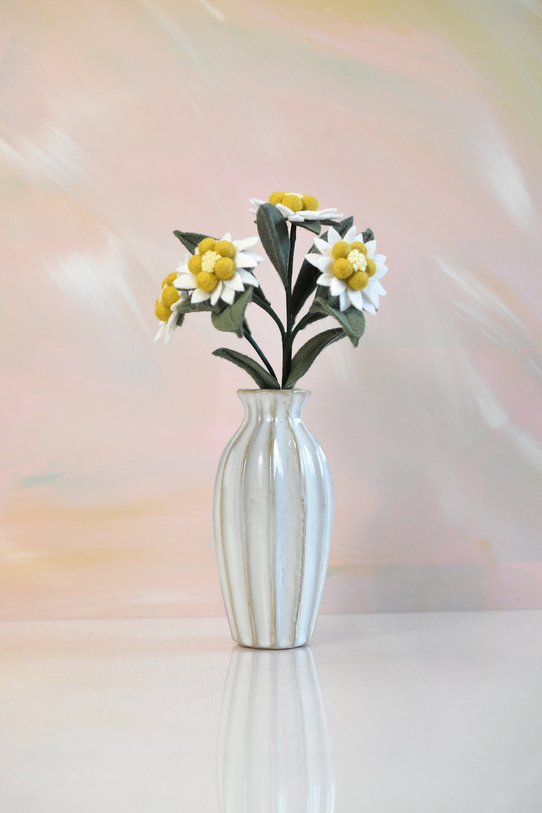 A white ribbed ceramic vase holding a bouquet of white daisies with yellow centers and green leaves, set against a soft pastel pink and peach blurred background.
