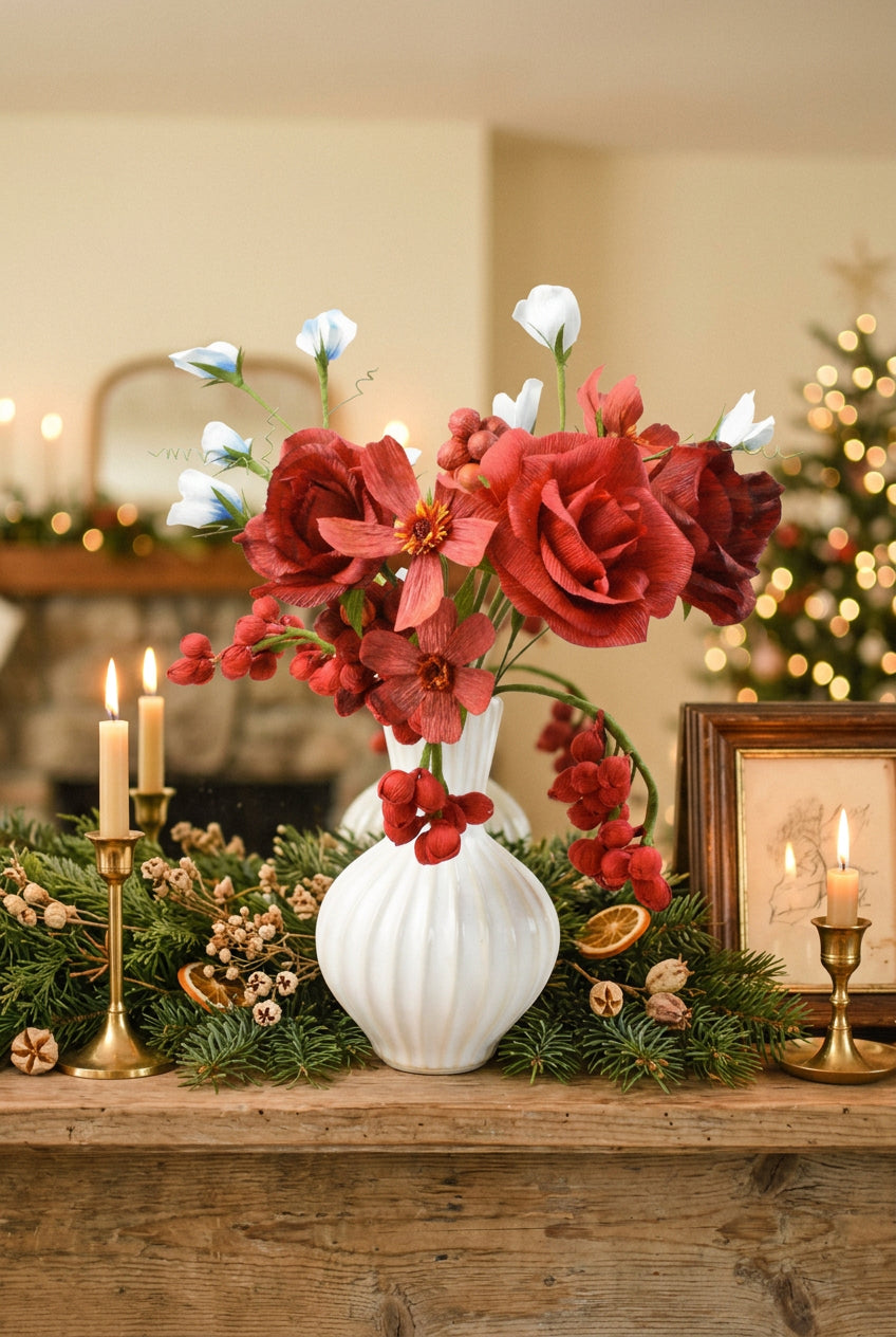 A floral arrangement featuring red roses, red clematis flowers, and small white flowers with green stems, presented in a white ribbed ceramic vase against a soft, light beige background.