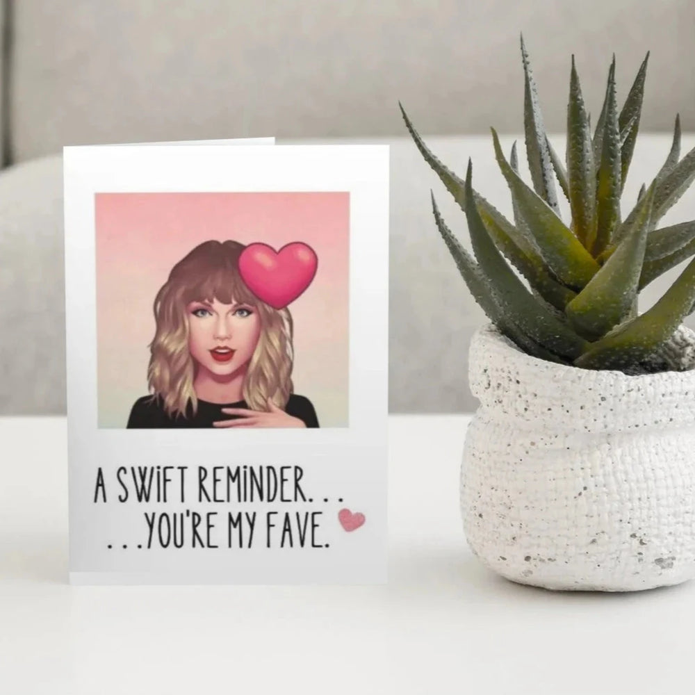 A greeting card with a colorful illustration of Taylor Swift on the front, alongside a white potted plant and a blank envelope.