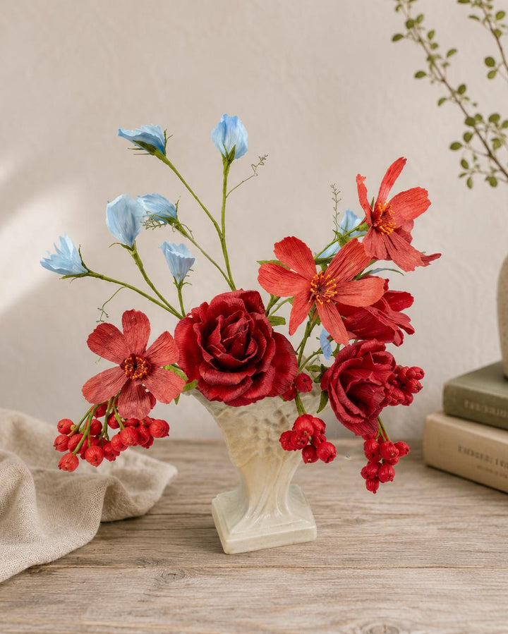 A floral arrangement in a small white vase on a wooden table, featuring red roses and flowers with light blue blossoms, styled beside a beige cloth and stacked books.