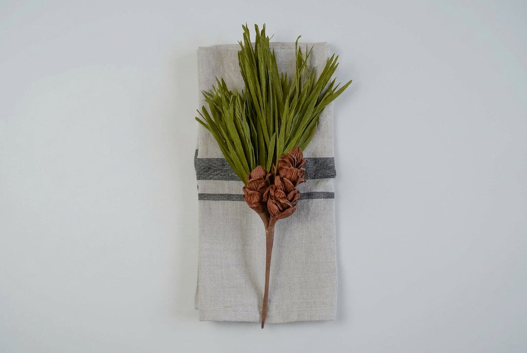 A rustic holiday table setting featuring a pine branch with green needles and brown pinecones wrapped in a white linen napkin with a gray stripe, placed on a gray wood plank background.