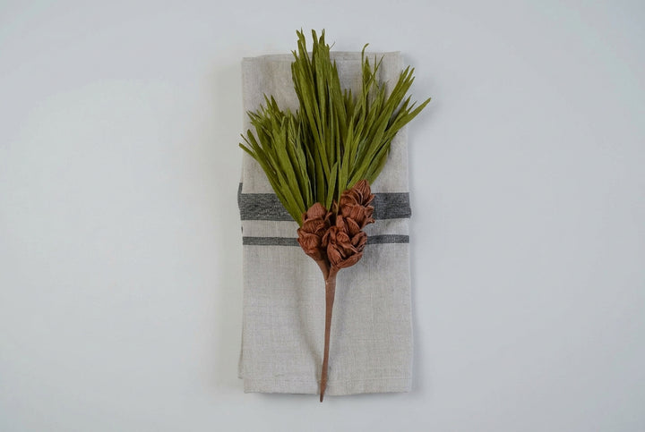 A rustic holiday table setting featuring a pine branch with green needles and brown pinecones wrapped in a white linen napkin with a gray stripe, placed on a gray wood plank background.