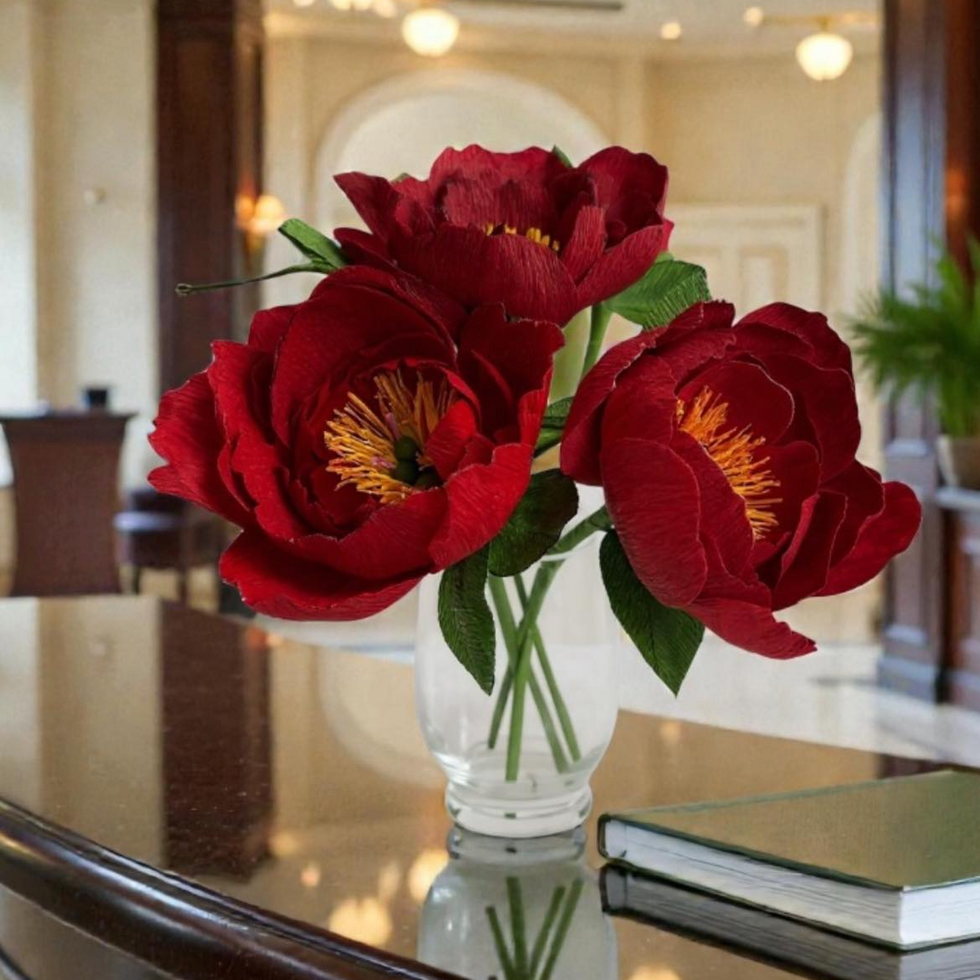 Three large artificial red peony flowers with yellow centers and green leaves arranged in a clear glass vase on a glossy dark table, with a blurred indoor room in the background.