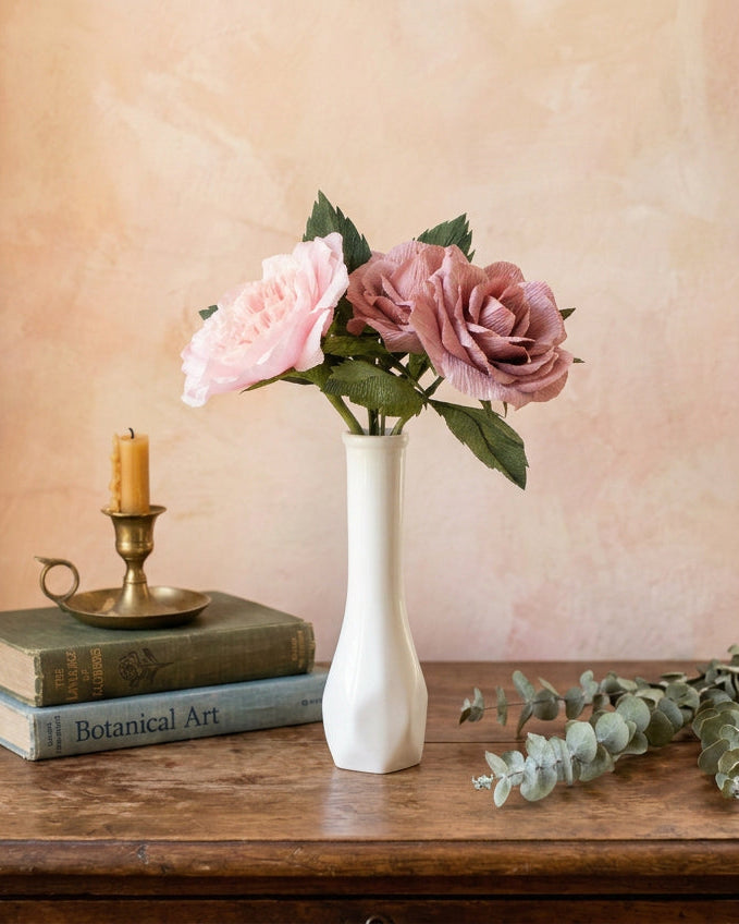 Two pink roses with green leaves in a small white ceramic vase, set on a white wooden surface against a soft pastel pink and yellow background.