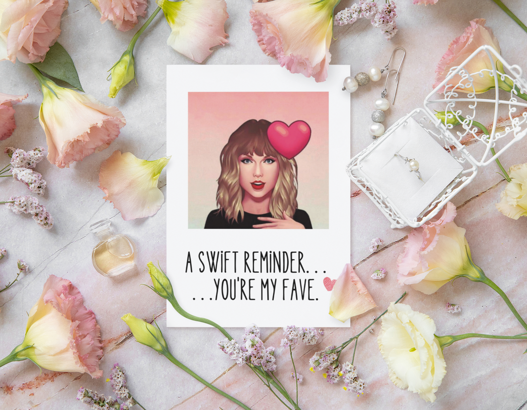 Card with a photo of  taylor swift and a heart, surrounded by flowers and jewelry on a marble surface.