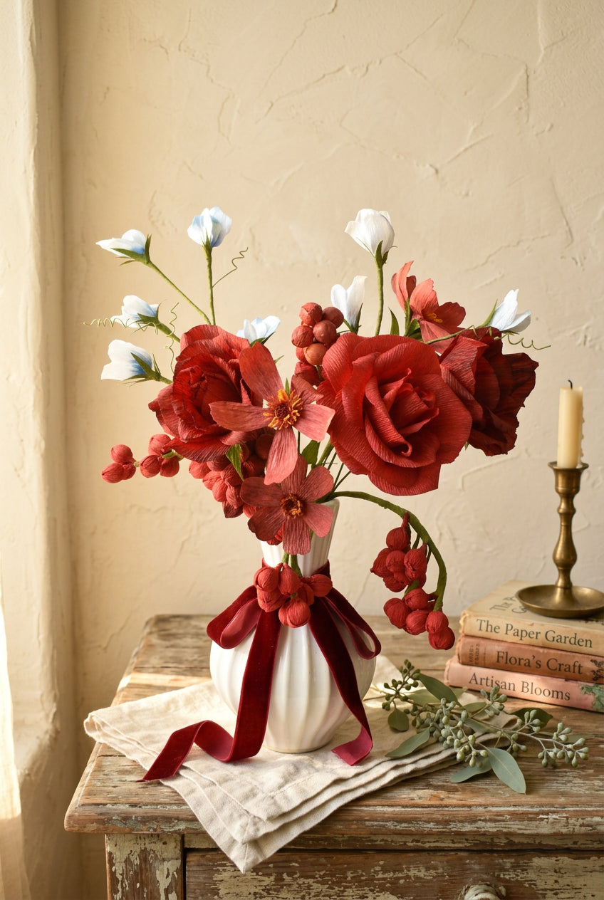 A floral arrangement featuring red and pink flowers with small white blossoms, displayed in a ribbed white ceramic vase against a soft pastel background.