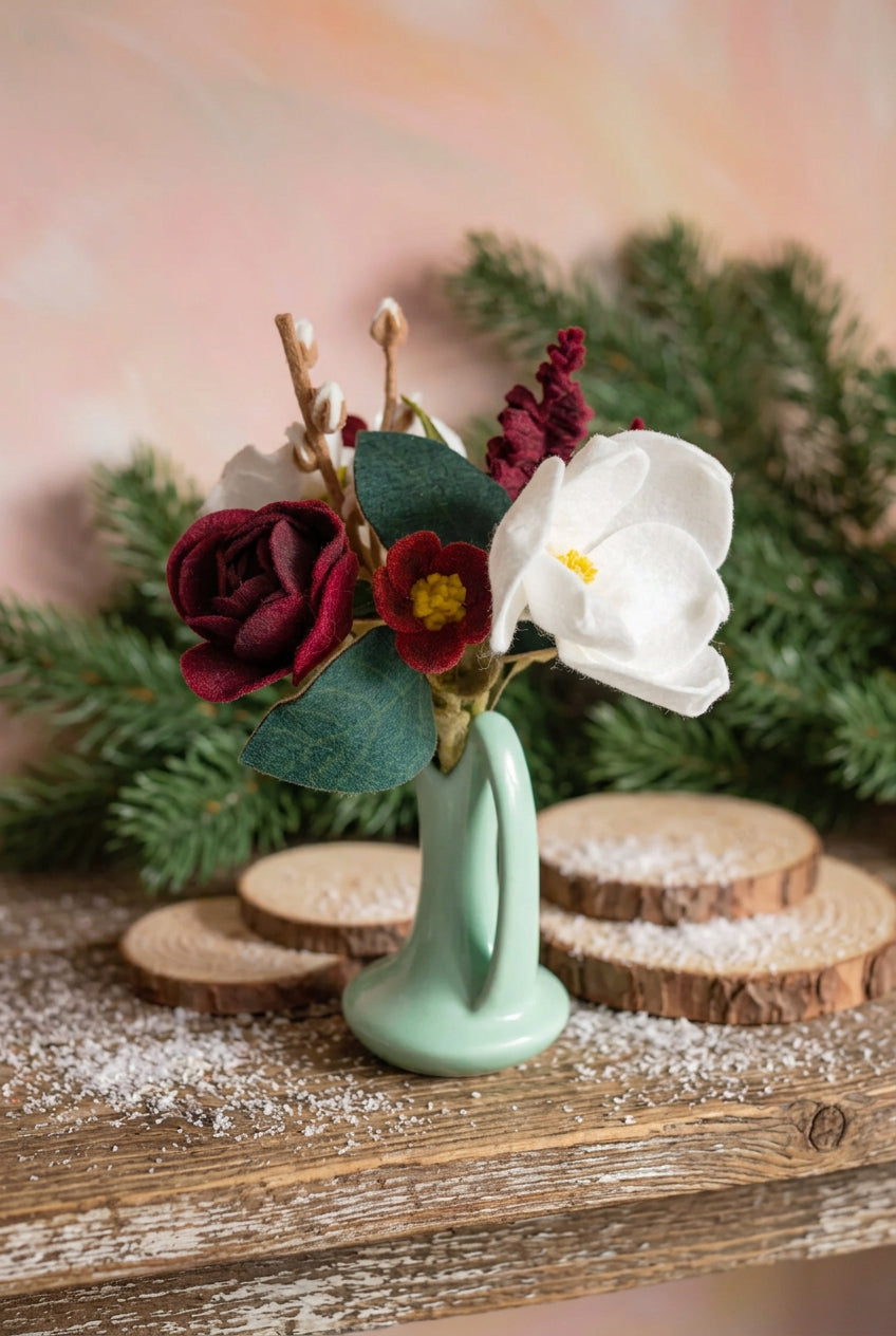 A small artificial floral arrangement in a pale mint green ceramic-style vase, featuring a large white flower, a deep burgundy rose, a burgundy flower with a yellow center, green leaves, and small white buds, set against a soft light pink and white background.